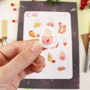 Op de afbeelding: Een vel stickers met verschillende winterse illustraties, waaronder een mok warme chocolademelk met marshmallows, een donut, een paar sokken en een stuk taart. De stickers zijn gedrukt op wit papier met een zwarte achtergrond.