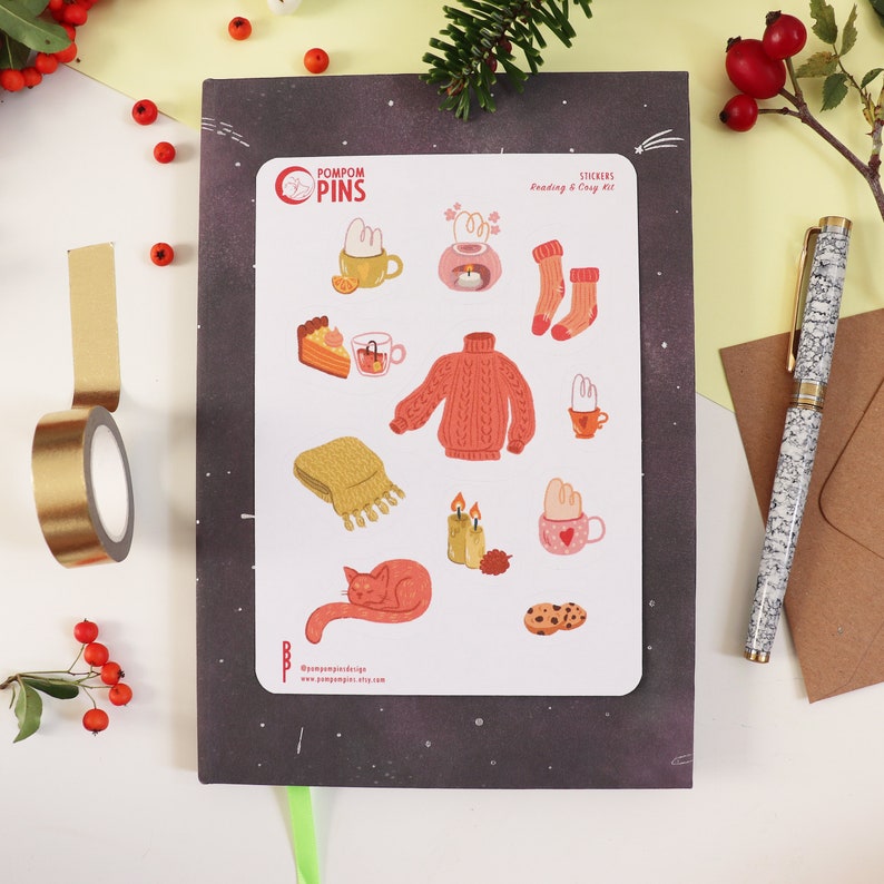 Op de afbeelding: Een vel stickers met een knus winterthema. De stickers bevatten illustraties van een kat, een trui, een mok warme chocolademelk, een sjaal, kaarsen, koekjes en meer. Het vel is gelabeld met "Pompom Pins" en "Stickers: Lezen & Gezellig Set".