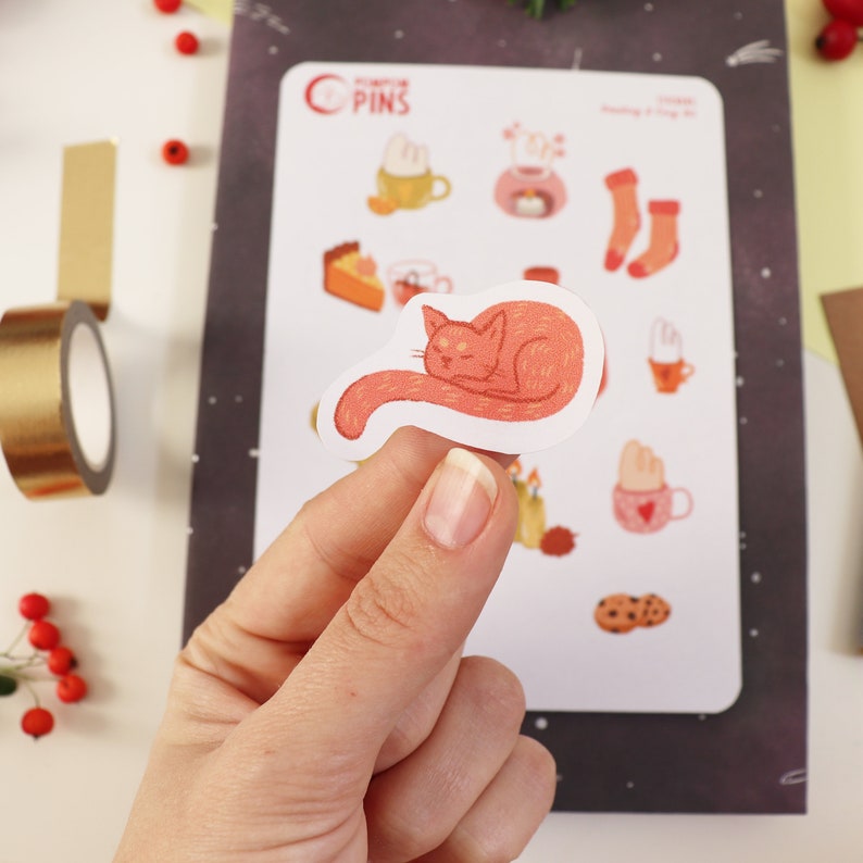 Op de afbeelding: Een vel stickers met verschillende schattige illustraties, waaronder een slapende kat, een mok warme chocolademelk, een stuk taart en een paar sokken. De stickers zijn gedrukt op wit papier en hebben een glanzende afwerking.