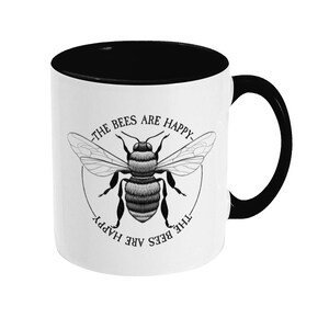May include: White ceramic mug with a black interior and handle. The mug features a black and white illustration of a bee with the text "The Bees Are Happy" on the front.