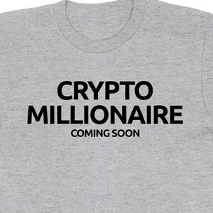 May include: A heather grey t-shirt with black text that reads "CRYPTO MILLIONAIRE COMING SOON".