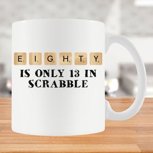 May include: White ceramic mug with a black graphic that reads "EIGHTY IS ONLY 13 IN SCRABBLE". The word "EIGHTY" is spelled out using Scrabble tiles.