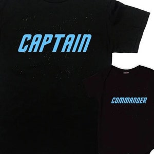 Puede incluir: Una camiseta negra con la palabra "CAPTAIN" en letras azules y un body de bebé negro con la palabra "COMMANDER" en letras azules. Ambas camisetas tienen un fondo estrellado en blanco y negro.