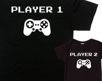 May include: Two black t-shirts with white text and a white video game controller graphic. The larger shirt says "Player 1" and the smaller shirt says "Player 2".
