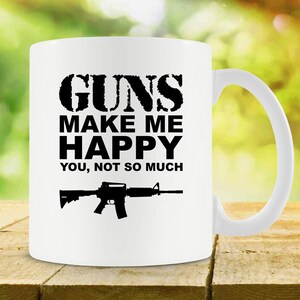 May include: White ceramic mug featuring the text "GUNS MAKE ME HAPPY YOU, NOT SO MUCH" in black, and a black rifle silhouette. The mug has a white handle. It is on a wooden surface.