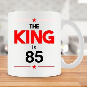 Puede incluir: Taza de café de cerámica blanca con un diseño rojo y negro. La taza dice "The King is 85" con una estrella roja a cada lado del texto.