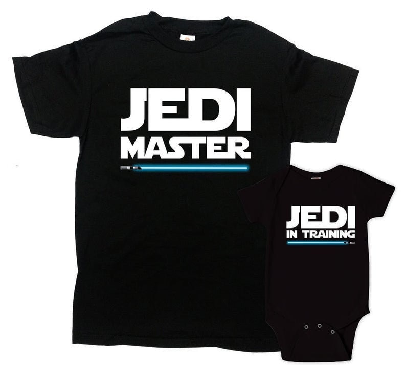 Op de afbeelding: Zwarte T-shirts met witte tekst. Het grotere shirt zegt "Jedi Master" met een blauwe lightsaber-afbeelding. Het kleinere shirt zegt "Jedi in Training" met een blauwe lightsaber-afbeelding.