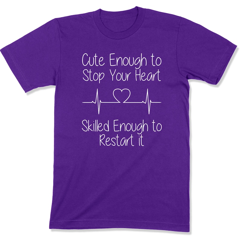 Puede incluir: Camiseta morada con texto blanco que dice "Cute Enough to Stop Your Heart" con una l&iacute;nea de latido del coraz&oacute;n y un s&iacute;mbolo de coraz&oacute;n. El texto contin&uacute;a con "Skilled Enough to Restart it".
