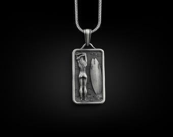 Può includere: Una collana con un ciondolo d'argento e un ciondolo rettangolare che raffigura una donna in piedi accanto a una tavola da surf. Il ciondolo ha una superficie testurizzata e un design in bianco e nero.