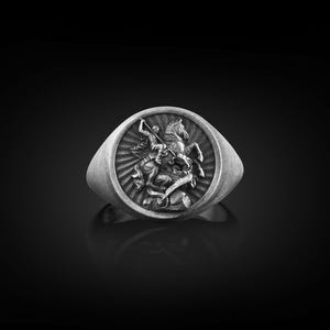 Puede incluir: Un anillo de plata con un dise&ntilde;o en relieve de un caballero a caballo matando a un drag&oacute;n.
