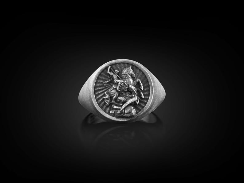 Puede incluir: Un anillo de plata con un dise&ntilde;o en relieve de un caballero a caballo matando a un drag&oacute;n.