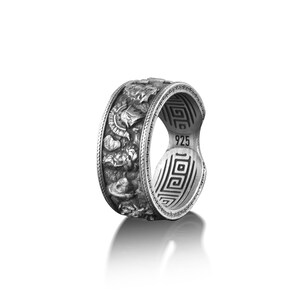 Pu&ograve; includere: Un anello d'argento con un designo dettagliato di guerrieri greci e un motivo a chiave greca sulla fascia. L'anello &egrave; contrassegnato "925" all'interno.