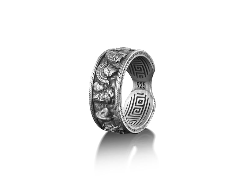 Pu&ograve; includere: Un anello d'argento con un designo dettagliato di guerrieri greci e un motivo a chiave greca sulla fascia. L'anello &egrave; contrassegnato "925" all'interno.