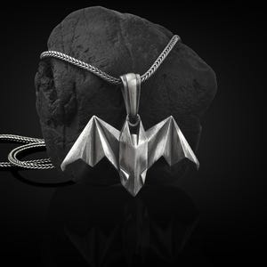May include: A silver origami bat pendant necklace with a braided chain. The pendant is hanging from a black rock.