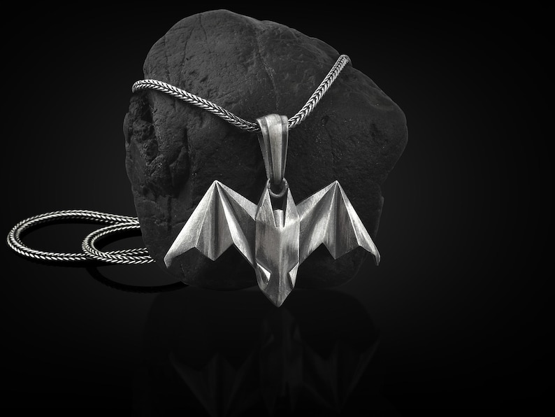 May include: A silver origami bat pendant necklace with a braided chain. The pendant is hanging from a black rock.