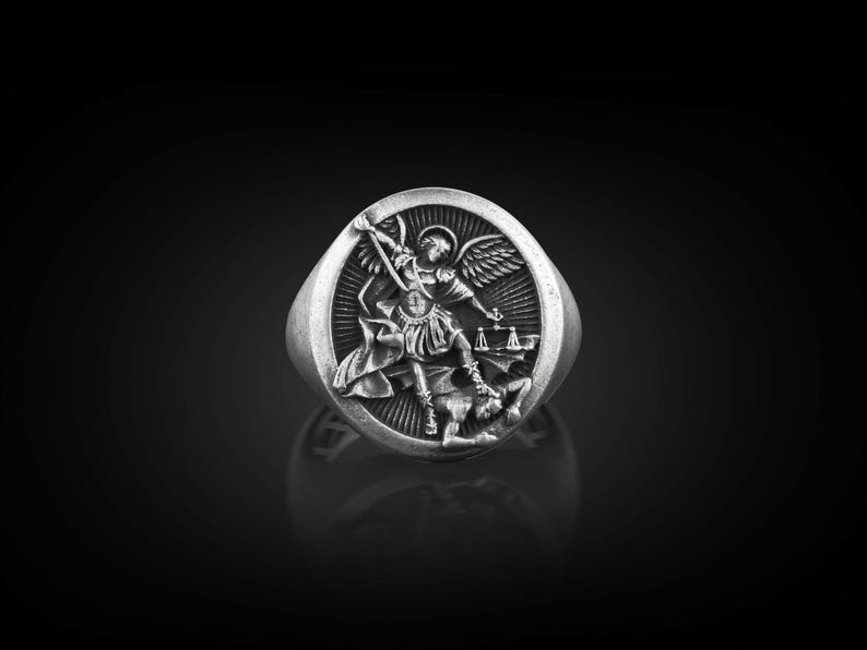 May include: A silver ring featuring an intricate design of an angel overcoming a demon. The angel is shown with wings and a sword, while the demon lies beneath the angel's foot.