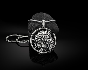 Puede incluir: Un collar de plata con un colgante redondo que presenta una representación detallada de la cara de un hombre con una corona de espinas. El colgante está sobre una superficie de roca negra.