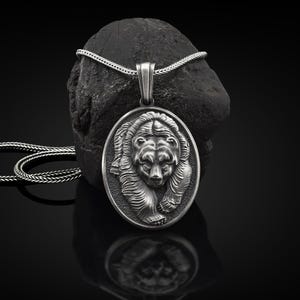 May include: A silver bear pendant necklace with a detailed, textured design. The pendant is oval-shaped and features a bear's head and shoulders. The necklace is on a silver chain.