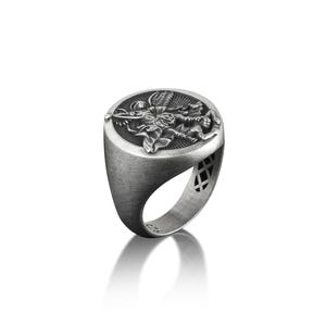 May include: A silver ring featuring an intricate design of an angel slaying a dragon. The ring is oval in shape and has a textured finish.