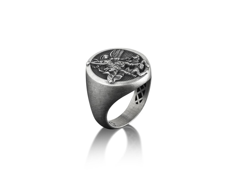 May include: A silver ring featuring an intricate design of an angel slaying a dragon. The ring is oval in shape and has a textured finish.