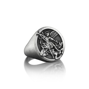 May include: A silver ring with a round face featuring a detailed depiction of an angel battling a demon. The angel is holding a sword and a scale, while the demon is lying on the ground.