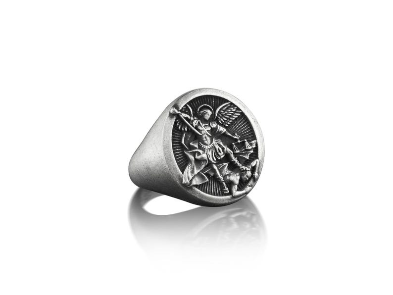 May include: A silver ring with a round face featuring a detailed depiction of an angel battling a demon. The angel is holding a sword and a scale, while the demon is lying on the ground.