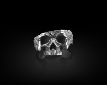 May include: A silver skull ring with a detailed design. The ring is on a black background.