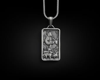 Puede incluir: Collar con colgante de plata que presenta un relieve detallado de un ángel con las alas extendidas, sosteniendo un pájaro y un ciervo, rodeado de otros animales. El colgante es rectangular y cuelga de una cadena de plata.