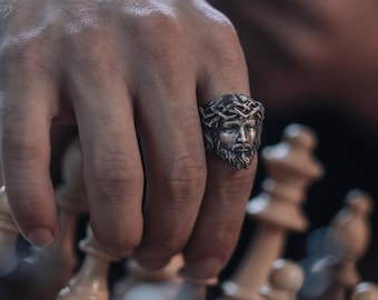 Op de afbeelding: Een zilveren ring met een gedetailleerde afbeelding van een mannengezicht dat een doornenkroon draagt. De ring wordt gedragen aan de wijsvinger van een hand.