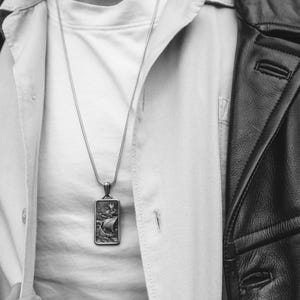 May include: A silver chain necklace with a rectangular pendant featuring a detailed design of a ship with a sail. The necklace is worn over a white shirt and a black leather jacket.
