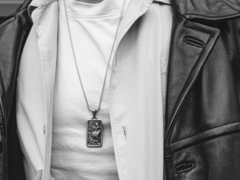 May include: A silver chain necklace with a rectangular pendant featuring a detailed design of a ship with a sail. The necklace is worn over a white shirt and a black leather jacket.