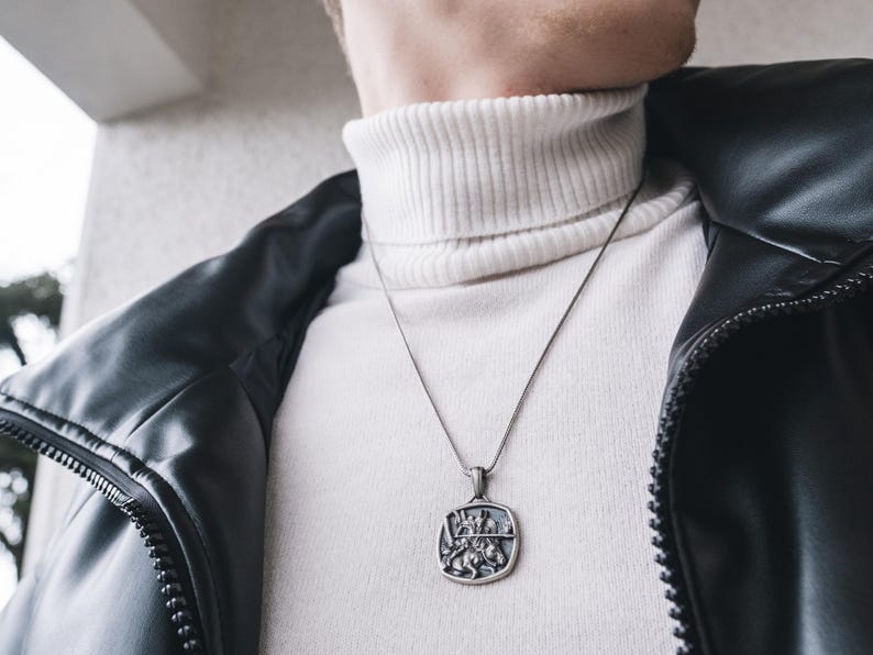 May include: A silver chain necklace with a square pendant featuring a detailed design of a knight on horseback. The necklace is worn by a person wearing a white turtleneck sweater and a black leather jacket.