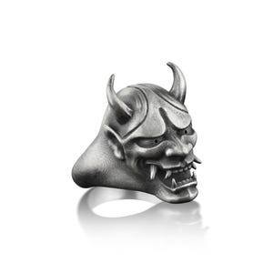 May include: A silver ring with a detailed oni mask design. The mask has sharp horns, a fierce expression, and a set of teeth.