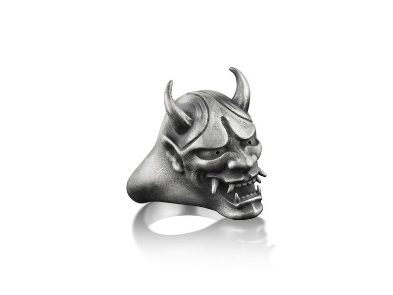 May include: A silver ring with a detailed oni mask design. The mask has sharp horns, a fierce expression, and a set of teeth.