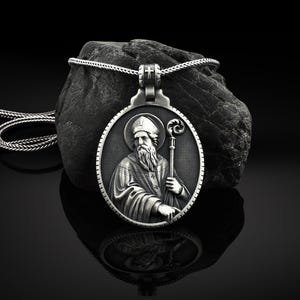 May include: Silver pendant necklace featuring a detailed relief of a religious figure holding a staff. The pendant is round and has a textured surface.