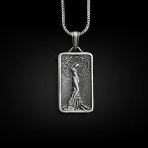 Puede incluir: Un collar de plata con un colgante rectangular que presenta una escultura en relieve detallada de una mujer de pie dentro de un árbol. El colgante está en una cadena de plata.