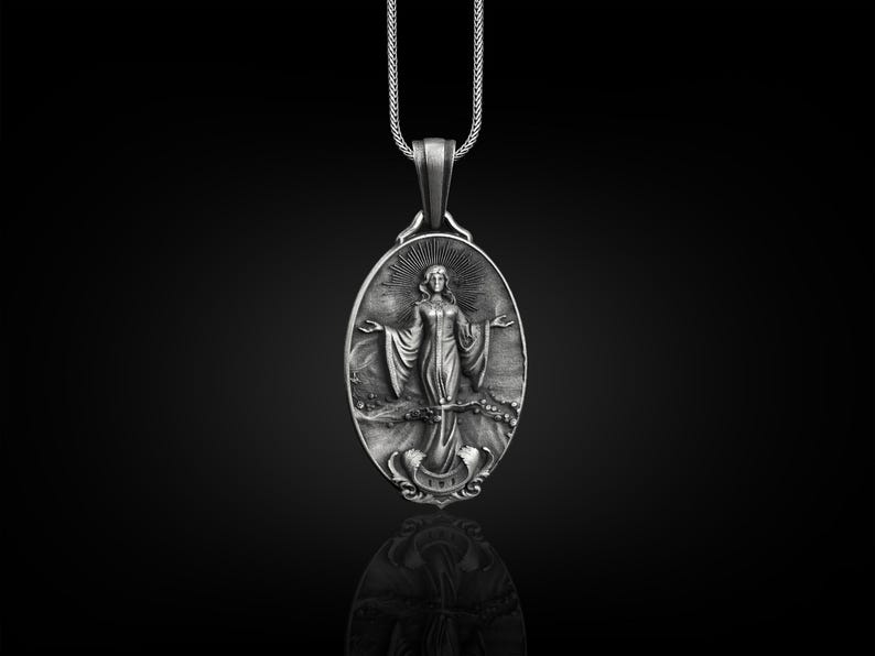 Puede incluir: Un collar con un colgante de plata que presenta un amuleto ovalado detallado con una imagen en relieve de una mujer de pie sobre una ola con un ancla debajo. El colgante est&aacute; en una cadena de plata.