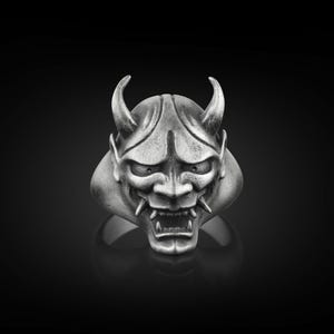 May include: A silver ring with a detailed design of a Japanese demon mask. The mask has sharp horns, a fierce expression, and a textured surface.