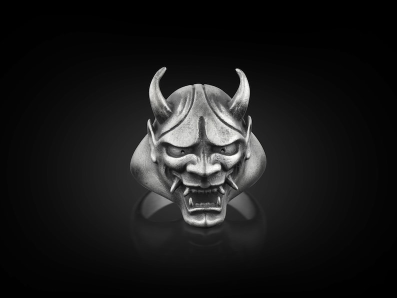 May include: A silver ring with a detailed design of a Japanese demon mask. The mask has sharp horns, a fierce expression, and a textured surface.