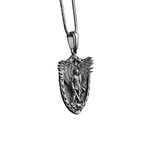 Puede incluir: Collar con colgante de plata que presenta un escudo con un &aacute;ngel que sostiene una espada. El &aacute;ngel tiene alas y est&aacute; representado en un dise&ntilde;o detallado en tres dimensiones.