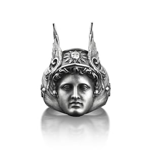 May include: A silver ring with a detailed design of a face wearing a winged helmet with a sun symbol on top.