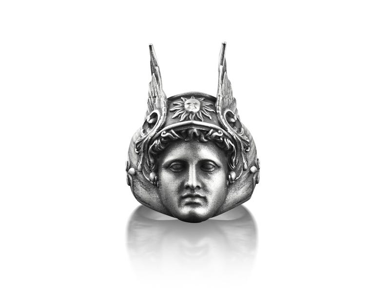 May include: A silver ring with a detailed design of a face wearing a winged helmet with a sun symbol on top.