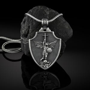 May include: Silver pendant necklace featuring a shield with an angel holding a sword and a cross. The angel is standing on a dragon. The pendant is on a black rock.