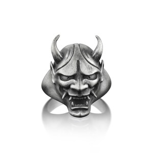 May include: A silver ring with a detailed oni mask design. The mask has sharp horns, a fierce expression, and a wide open mouth.