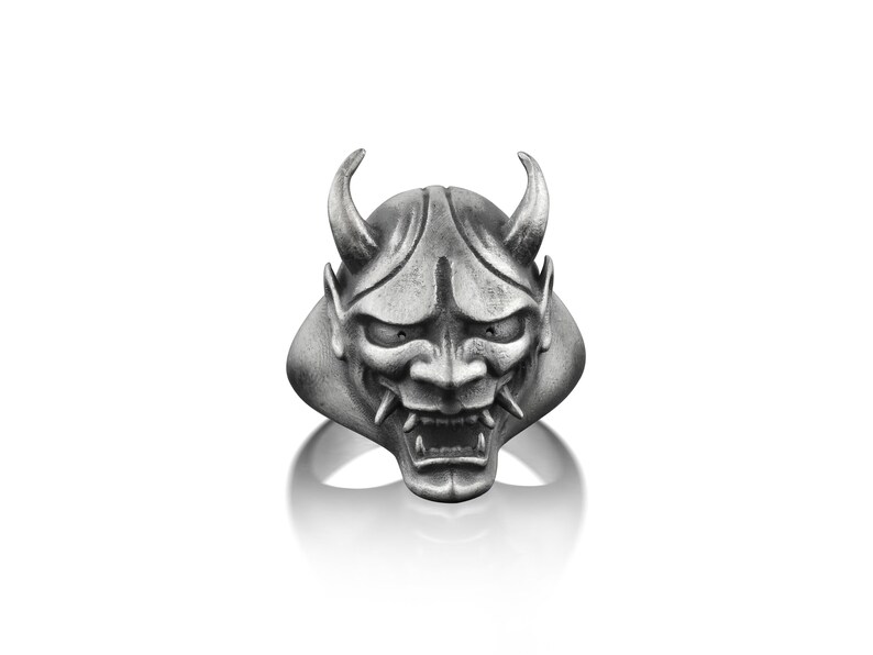 May include: A silver ring with a detailed oni mask design. The mask has sharp horns, a fierce expression, and a wide open mouth.