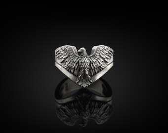 Può includere: Anello color argento con un design dettagliato di un'aquila con le ali spiegate. L'aquila è il punto focale, con intricati dettagli delle piume. L'anello è posto su uno sfondo scuro, evidenziando la lucentezza metallica.