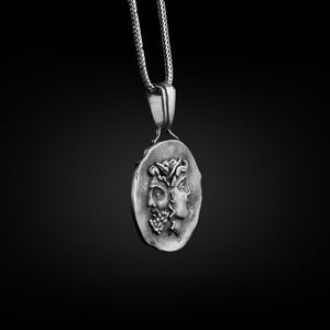 May include: A silver necklace with a pendant featuring two faces in profile. The pendant is round and has a textured surface.