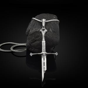 May include: A silver sword pendant necklace with a black braided chain. The sword has a detailed design and the inscription "FOR - A - MAN" on the blade.
