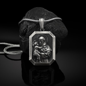 Op de afbeelding: Een zilveren ketting met een rechthoekige hanger met een gedetailleerd beeld van Sint Antonius van Padua die een baby Jezus vasthoudt. De hanger heeft een zwarte achtergrond en ligt op een zwarte ondergrond.