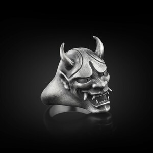 May include: A silver ring with a detailed oni mask design. The mask has horns, sharp teeth, and a fierce expression.